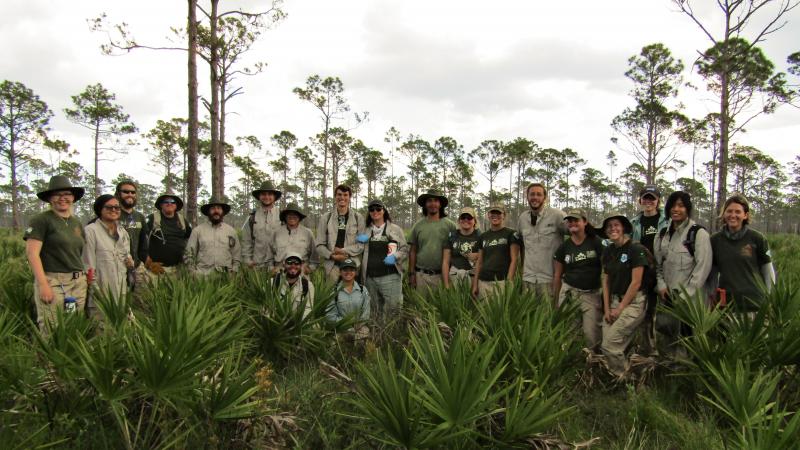 A group of FLCC members pose in a pine scrub landscape, wearing khaki and green uniforms and smiling. 
